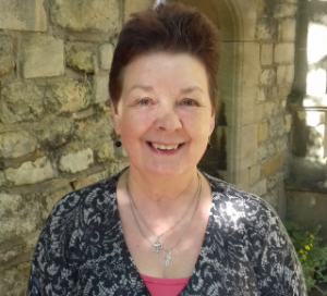 Rev. Sue Woolley wearing a pink t-shirt, grey cardigan and chalice necklace, standing in front of a stone building.