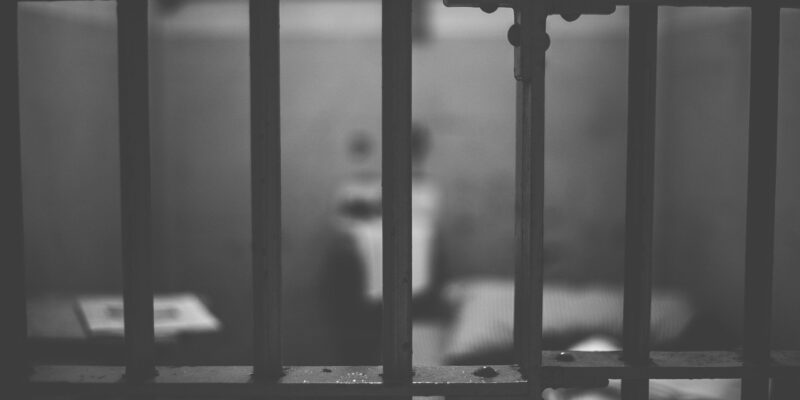 A black and white image of a prison cell seen through the bars.