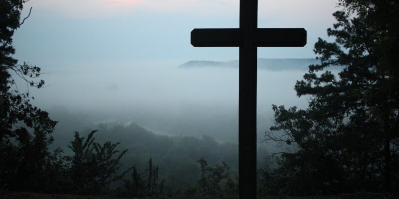 A wooden cross silhouetted against a misty wooded hillside.